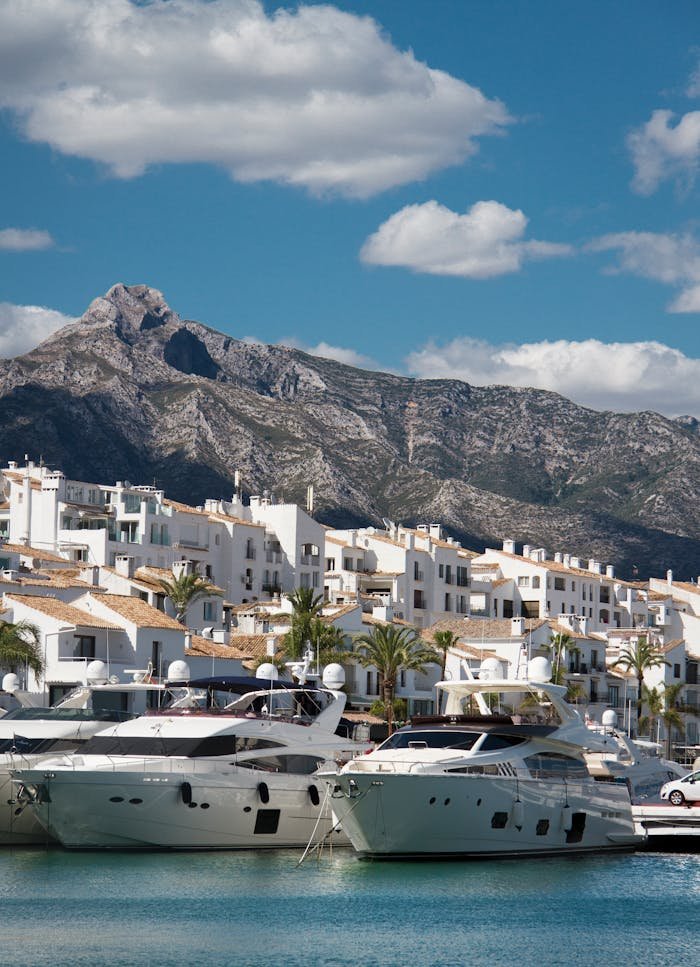 Elegant yachts docked at a Mediterranean harbor with picturesque mountain and town backdrop.