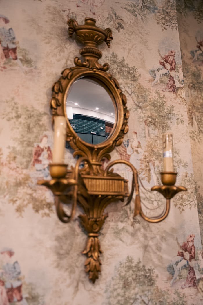 Elegant ornate mirror with attached candle holders on floral wallpaper, adding a classic touch.